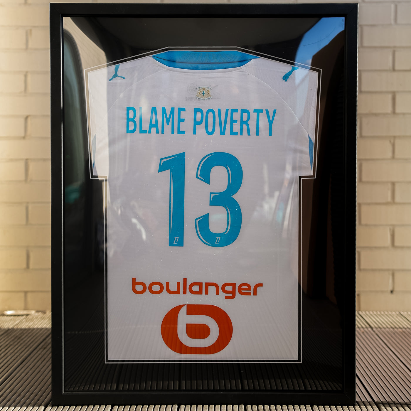 Black framed white football shirt personalised with BLAME POVERTY and the number 13, against a white brick wall