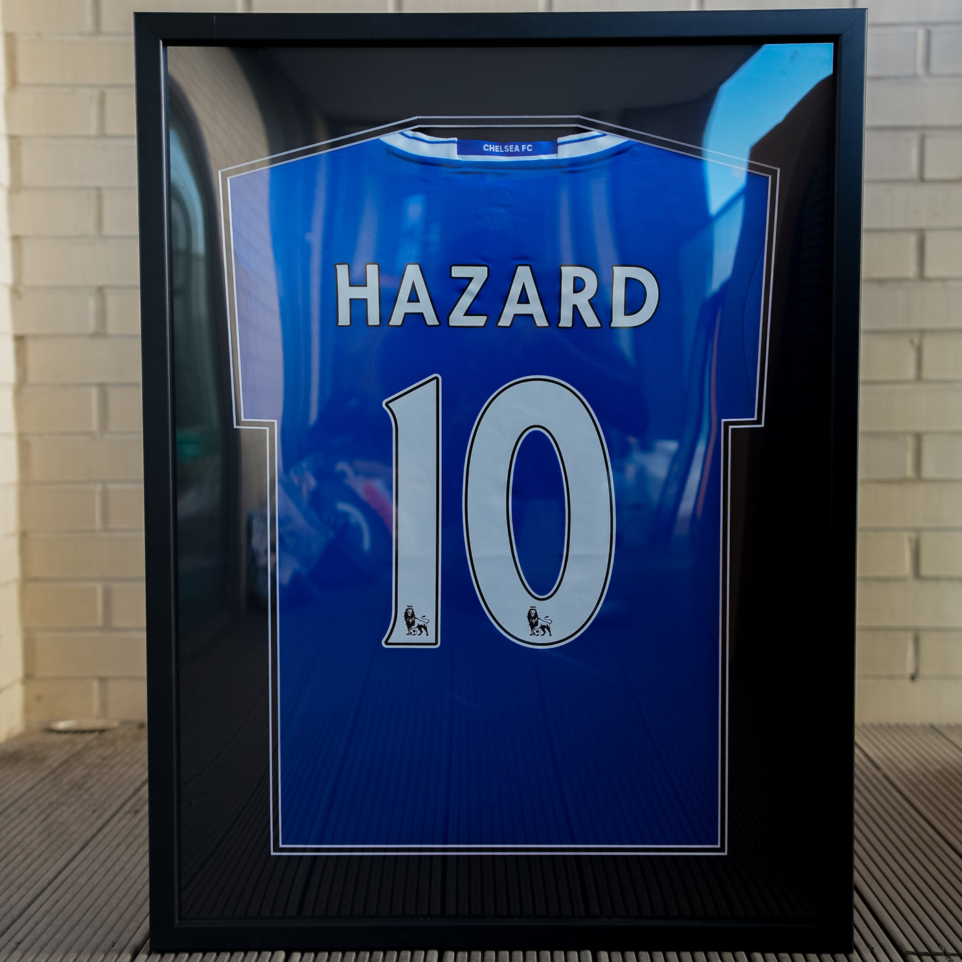 Black framed blue football shirt personalised with a name and the number 10 in white lettering, against a white brick wall