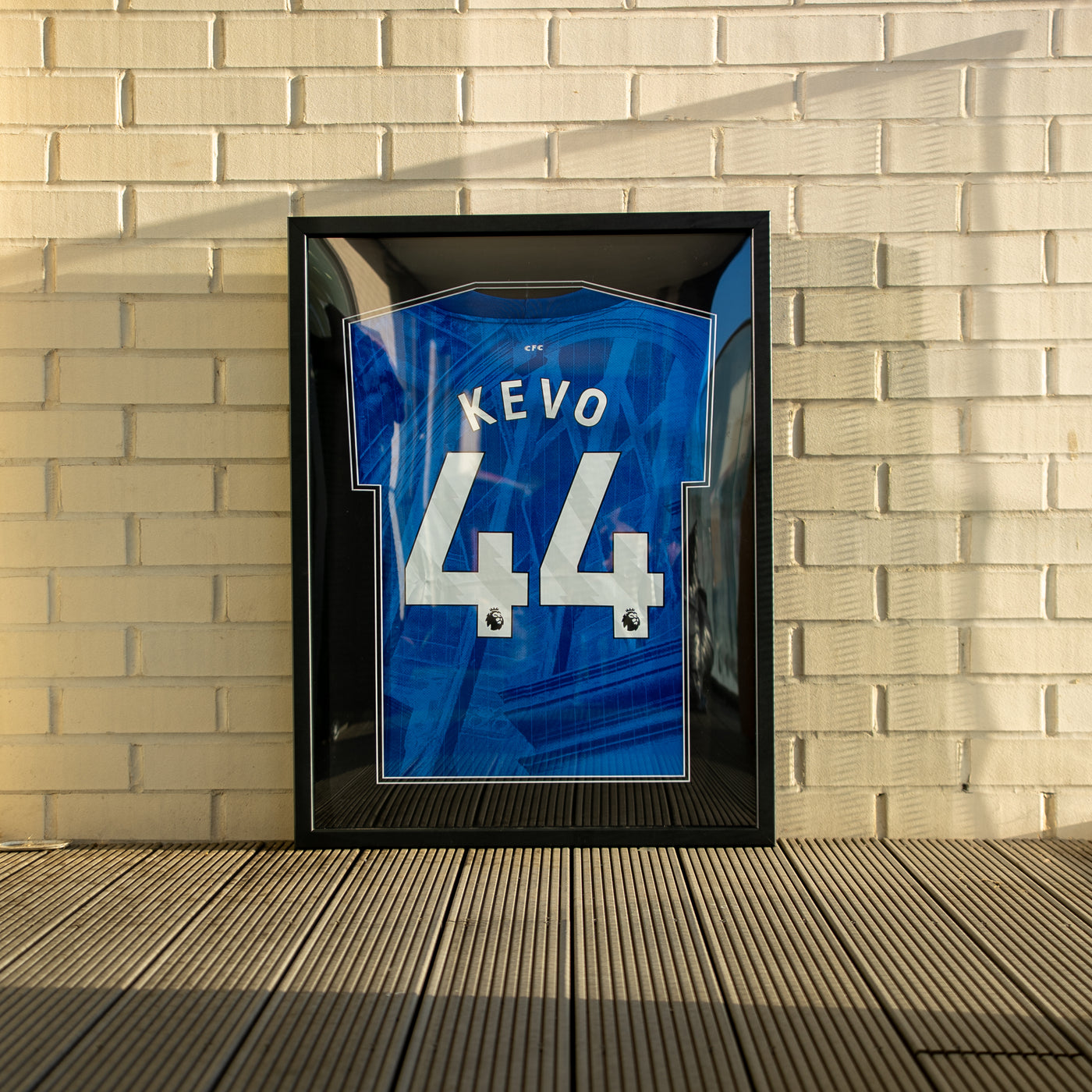 Black framed white football shirt personalised with BLAME POVERTY and the number 13, against a white brick wall